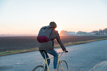 Fototapeta premium Female commuter riding a bike out of town to a suburban area. Young woman goes home by bike from work along the road at sunset
