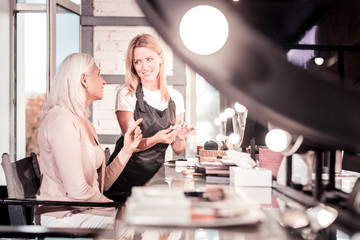 Friendly conversation. Cheerful beautician keeping smile on her face and holding cosmetics while looking at her client