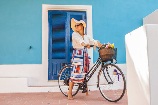 Beautiful young girl with bicycle in front of a blue house wearing colorful fashion dress with large hat in Sunny summer day, in Ponza island Italy
