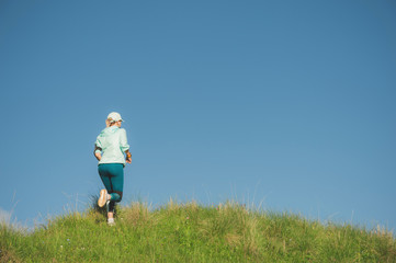 Naklejka na ściany i meble Attractive sports girl in a cap and headphones jogging through the grass in a picturesque place up the hill against the blue sky. Workout outdoors