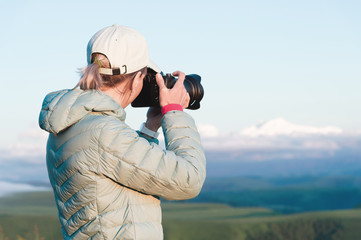 Obraz premium Portrait of a girl photographer in a cap on nature photographing on her digital mirror camera. Back view