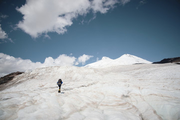 Traveler in a cap and sunglasses with a backpack on his shoulders in the snowy mountains on the glacier against the sky and clouds. Traveler in a natural environment