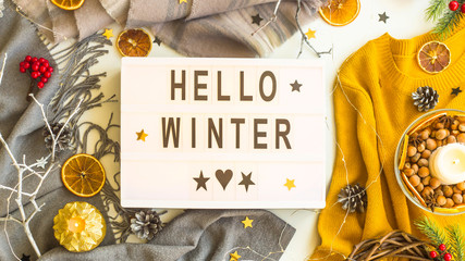 Hello winter. Written in black letters on a rectangular decorative lamp among winter women's sweaters and decor - candles, oranges, stars. Top view, flat lay