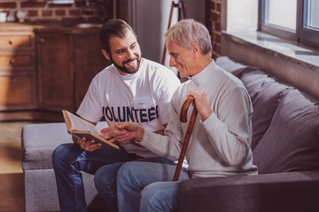 Avid readers. Glad bearded volunteer reading a book for the aged man and smiling