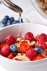Healthy breakfast with fresh honey is pured into the flakes, muesli, raspberries, blueberries, strawberries on a gray wooden table. Close-up.