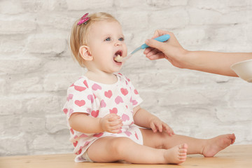 Mother feeding her baby with a spoon, close up portrait. Adorable blonde baby girl eating a healthy prepared meal with mother help at home