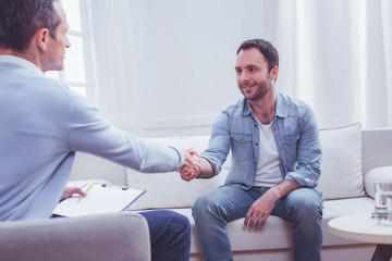 Gratitude. Pleased positive bearded man shaking hands with professional psychiatrist while being extremely thankful to him for a high quality assistance - obrazy, fototapety, plakaty