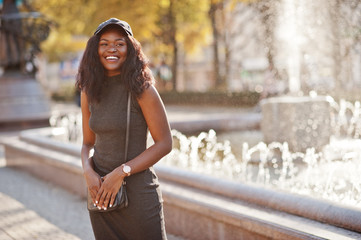 Stylish african american girl in gray tunic, crossbody bag and cap posed at sunny autumn day...