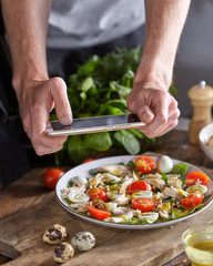Appetizing healthy salad from organic ingredients in a plate with mens hand making the photo by smartphon on a wooden table.