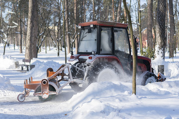 a small tractor with a rotating brush clears a wide road in the city park from the fresh fallen...