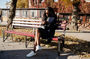 Naklejka premium Stylish african american girl in blue coat and sunglasses posed at sunny autumn day at bench and using her mobile phone. Africa model woman with smartphone.