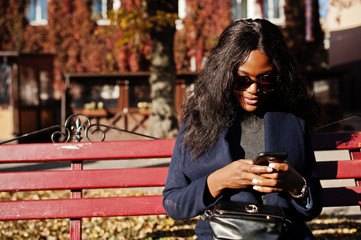 Stylish african american girl in blue coat and sunglasses posed at sunny autumn day at bench and using her mobile phone. Africa model woman with smartphone. - obrazy, fototapety, plakaty