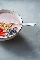 White bowl with homemade granola, sliced strawberries, yogurt, blueberry, spoon - natural breakfast on a gray table.