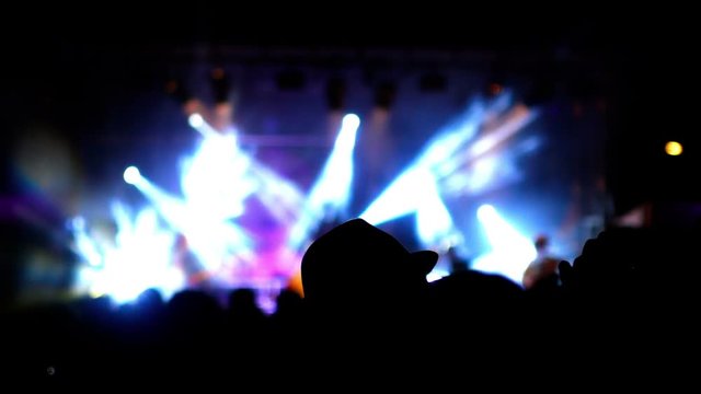 Blurred musicians and lighting effects on a rock concert stage at night. A man with a hat and the crowd stand in shadow in foreground.