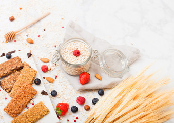 Organic cereal granola bar with berries on marble board with honey spoon and jar of oats and linen towel on marble background. Raw wheat bundle