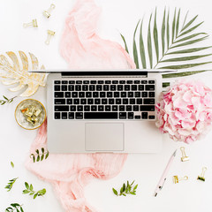 Modern home office desk workspace with laptop, pink hydrangea flowers bouquet, tropical palm leaf, pastel pink blanket, monstera leaf plate and accessories on white background. Flat lay, top view.