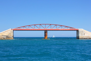 Small white sailboat ship in blue water of Grand Harbor, Valletta. Red Breakwater Bridge metallic construction on foreground