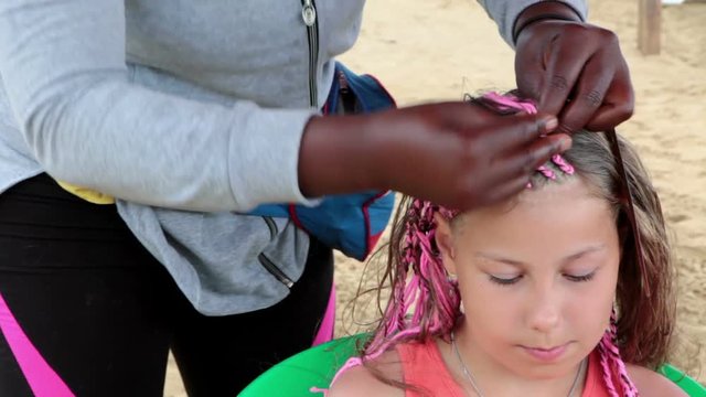 Interweaving pink threads into braids. Afro American woman makes dreadlocks for European girl
