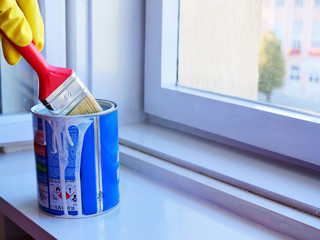 Hand in yellow rubber gloves dips a paintbrush in paint can to load it with white paint. Window frame restoration in home interior.