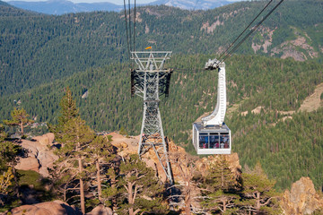 A cable car gondola moves toward a cable support structure to hold up the cables. The cable car holds many people. The cable structure stands on rugged mountain peak. © Timothy