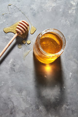 Wooden stick on a spilled honey on the table and glass pot with natural meadow sweet dessert on a gray kitchen table. Pure natural sweet goodness.