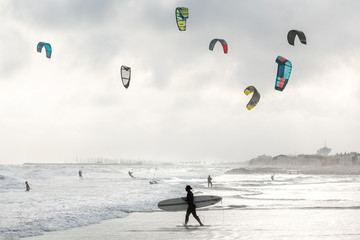 un surfer passe devant des kitesurf sous un ciel nuageux - obrazy, fototapety, plakaty