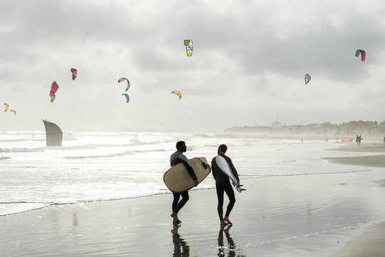 deux surfers marchant sur une plage avec des voiles de kitesurf en fond sous un ciel nuageux