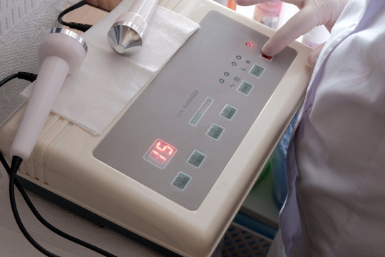 Close-up of a white-gloved hand pressing a power button on a digital ultrasonic massager device for skin. The concept of cosmetic or medical subjects.