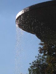 Plato de fuente ornamental  de piedra con gotas de agua cayendo y árbol de fondo con cielo azul