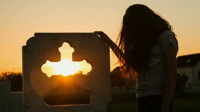 Young lady grieving near a funeral cross at sunset