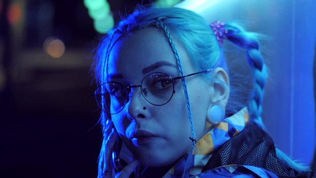 Attractive pretty girl with unusual hairstyle near glowing neon lights of the city at night talking with smartphone. Dyed blue hair in braids. Happy hipster teenager using mobile