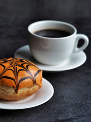 Orange halloween donut decorated with chocolate spider web and white cup of coffee on dark background.