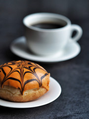 Orange halloween donut decorated with chocolate spider web and white cup of coffee on dark background.
