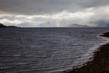 Scenic Scottish landscape with a silver grey lake and visible shore as well as cloudy sky with sunny spells shining onto green mountains in background