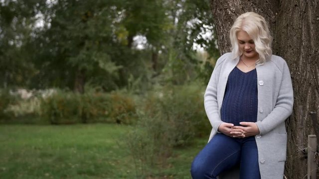 HD Pregnant woman standing next to tha tree and looks at her belly