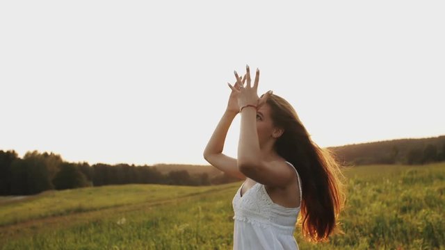 Happy young woman in white dress smiling, laughing and showing up in the sky with her hands standing in the field in the sunlight at sunset on a summer evening, pretty girl in a good mood