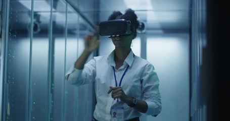 Formal African American woman wearing VR headset and working in corridor of data center among server racks - Powered by Adobe