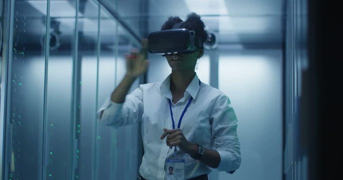 Formal African American woman wearing VR headset and working in corridor of data center among server racks