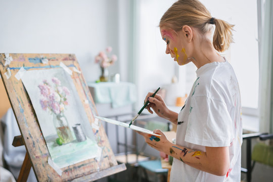 A young artist girl with a face and clothes in the paint, paints a picture on the easel.