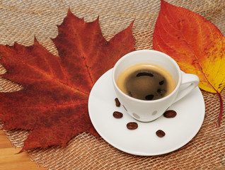 A cup of black coffee and coffee beans with canvas and red leaves as background