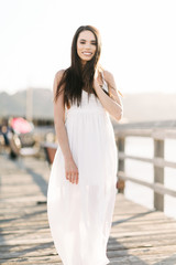 Portrait of a smiling young woman in a white summer dress standing on a pier during the golden hour sunset.