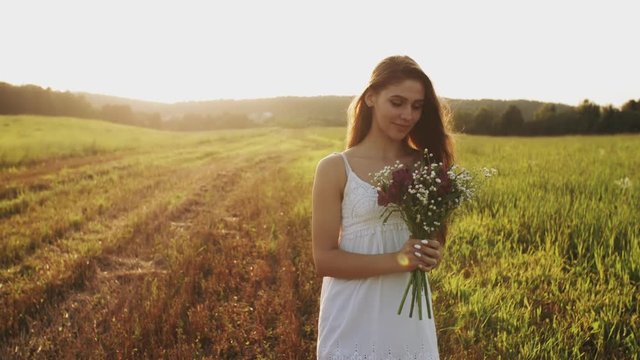 Young woman in white dress standing in a meadow with bouquet of wild flowers on summer evening. Long hair flowing in wind in sunshine at sunset, she looks up into the camera and smiles