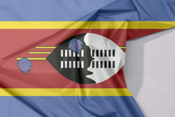 Fototapeta na wymiar Swaziland fabric flag crepe and crease with white space, blue yellow and red with the large black and white Nguni shield and the staff decorated with the feather tassels.