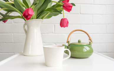White cup on small table with green teapot and red tulips in jug at rear (selective focus)
