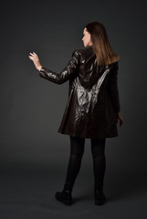 full length portrait of brunette woman wearing long leather coat. standing pose  with back to the camera, on grey studio background.