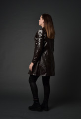 Fototapeta na wymiar full length portrait of brunette woman wearing long leather coat. standing pose with back to the camera, on grey studio background.