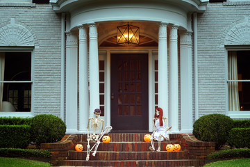The house is decorated for Halloween: Two skeletons with orange pumpkins sitting near the entrance to the house. Evening