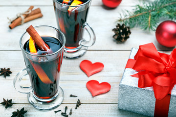 Two glasses of hot mulled wine with cinnamon, oranges, anise and gift box, hearts on wooden table. 
