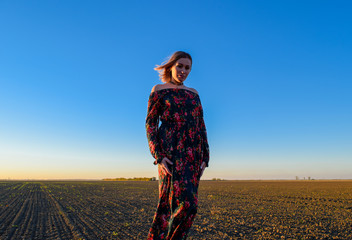 Woman in a plowed field in a red-black dress on a sunset background