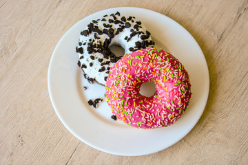 An appetizing donut with pink icing and a multi-colored sprinkle and a delicious chocolate donut with white icing sprinkled with chocolate chips and on a white plate
